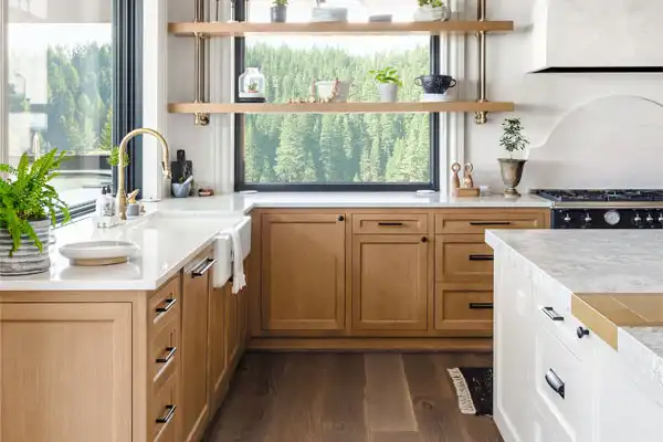 Modern kitchen with wooden cabinets, white countertops, and gold faucet. Open shelving with plants and dishes. Large window overlooks a forested area.