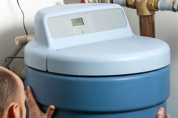 Person adjusting a blue water softener unit with a digital display, installed next to plumbing pipes.