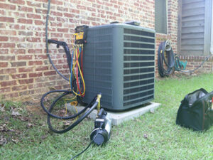 An air conditioning unit installed next to a brick wall, with equipment and hoses attached and a toolbox placed nearby on the grass.