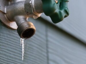 Close-up of an outdoor water tap with a green handle, showing thin icicles hanging from the spout against a house's siding.