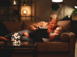 A person reads a book to a child on a cozy sofa in a dimly lit room with warm lighting and a decorative table in the foreground.