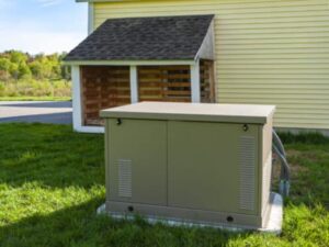 A large gray electrical generator is installed on a concrete pad in the grass near a yellow building with a small attached structure.