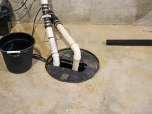 Basement sump pump with PVC pipes, a black lid, and a bucket nearby, set against a concrete wall and floor.