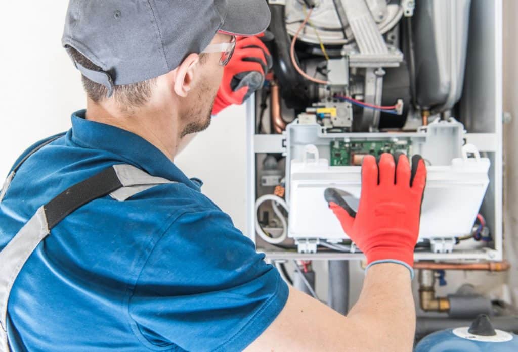 An HVAC technician wearing protective gloves inspects and repairs the electrical components inside a residential furnace.